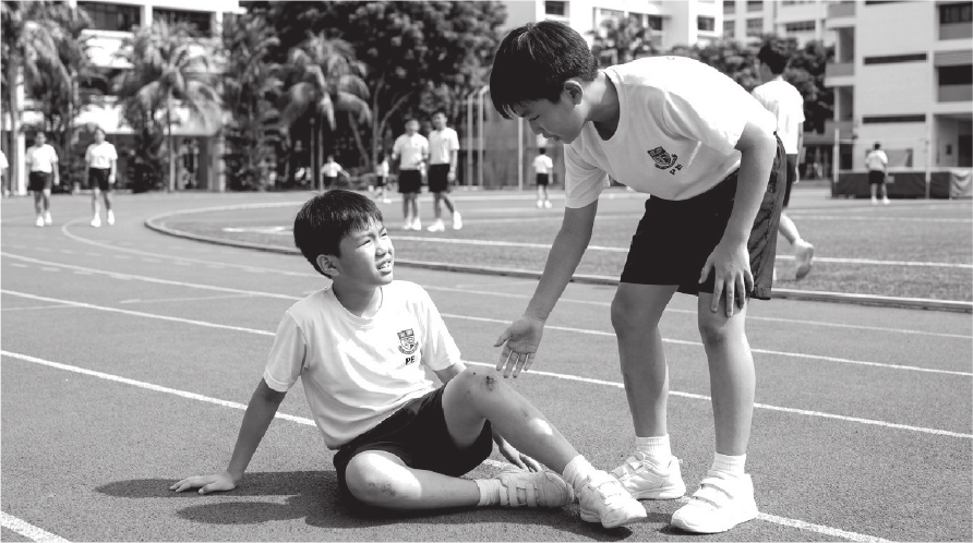 A boy on a running track bending down to help his friend who has fallen and scraped his knee.